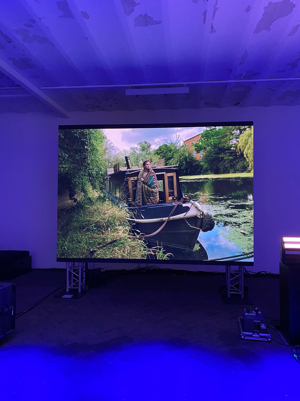 A Life at Home portrait of a woman on a canal narrowboat shown on a large screen in a blue-lit gallery