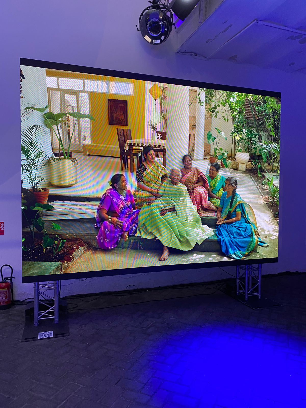 A large screen in a blue-lit room shows a Life at Home portrait of women in saris gathered on a veranda in India