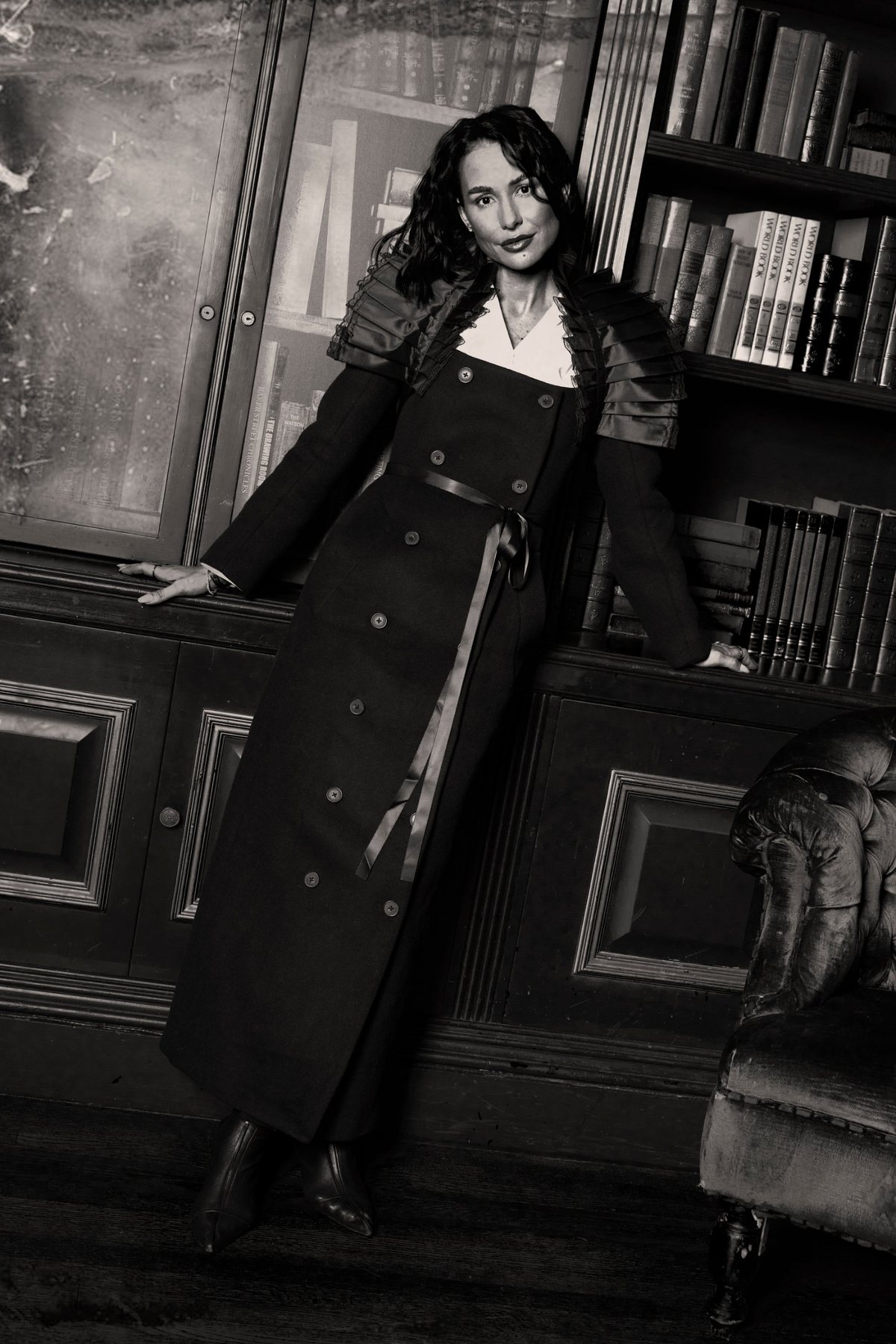 A creator in a long coat dress with pleated shoulders, leaning against the library shelves